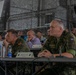 A multinational military conference or briefing is taking place in what appears to be a military hangar or field facility decorated with camouflage netting. Several senior military officers from different nations, including Spain and the Czech Republic, are seated at tables alongside civilian officials. Name placards are visible on the tables, identifying at least one participant as Maj Gen Petr Svoboda, Territorial Forces Command Commander.