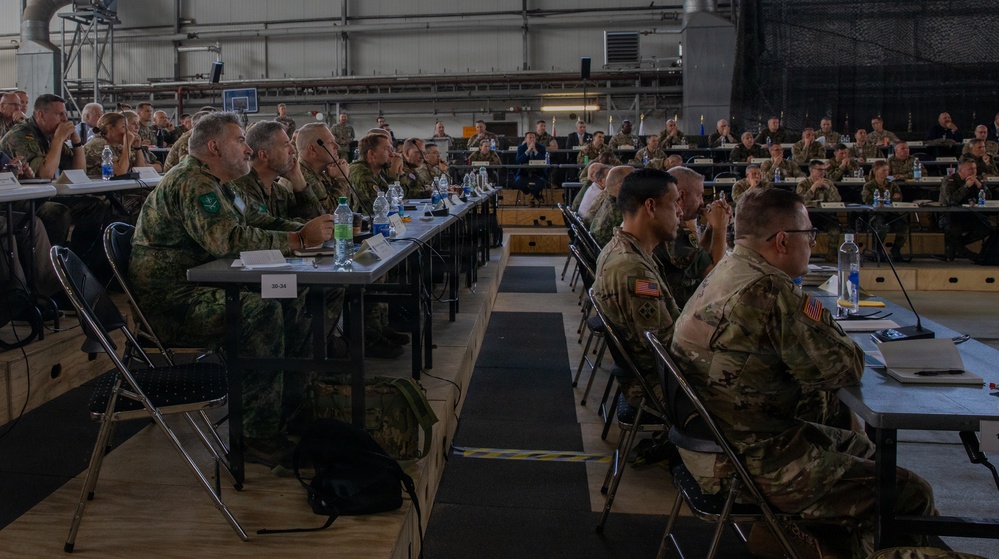 A large military briefing or conference is taking place inside what appears to be a hangar or large indoor facility. Numerous military personnel from multiple nations, wearing various camouflage uniforms, are seated at long tables arranged in rows facing a common direction. The gathering suggests a multinational military exercise, summit, or joint operations planning session.