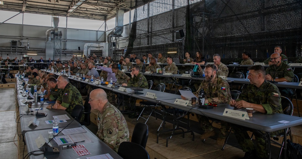 A large multinational military conference or briefing is taking place inside what appears to be a military hangar or large industrial facility. Numerous military personnel in various camouflage uniforms are seated at long rows of tables, suggesting a coalition or allied forces gathering. Name placards are visible on the tables, indicating a formal, organized meeting structure.