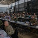 A large multinational military conference or briefing is taking place inside what appears to be a military hangar or large industrial facility. Numerous military personnel in various camouflage uniforms are seated at long rows of tables, suggesting a coalition or allied forces gathering. Name placards are visible on the tables, indicating a formal, organized meeting structure.