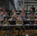 A formal military briefing or conference is taking place inside what appears to be a large hangar or industrial facility. Multiple rows of military personnel in various camouflage uniforms, along with a few civilians, are seated at long tables facing a presenter or screen outside the frame. The setting includes name placards, laptops, water bottles, and documents, indicating an organized multi-national or joint military meeting.
