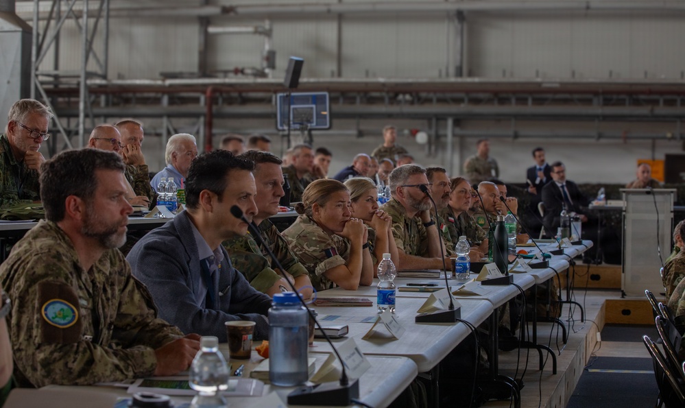 A multinational military briefing or conference is taking place inside what appears to be a large hangar or industrial facility. Personnel in various military uniforms and civilian attire are seated at long tables arranged in rows, facing a presentation area. The gathering includes both military officers and civilian officials, suggesting a joint or coalition-level meeting.