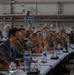 A multinational military briefing or conference is taking place inside what appears to be a large hangar or industrial facility. Personnel in various military uniforms and civilian attire are seated at long tables arranged in rows, facing a presentation area. The gathering includes both military officers and civilian officials, suggesting a joint or coalition-level meeting.