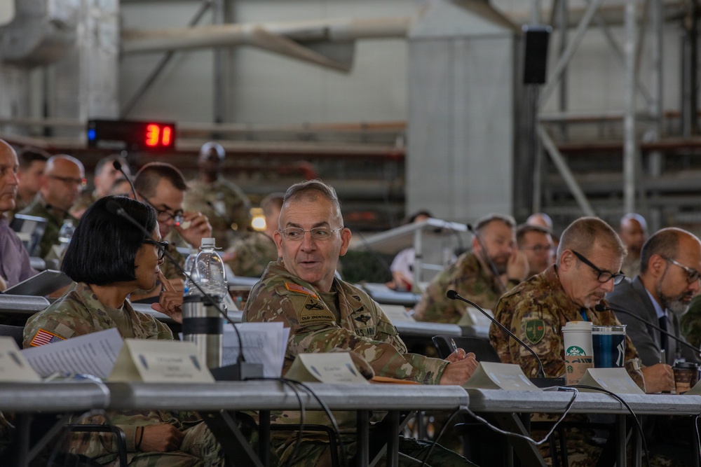 A military briefing or conference is taking place in what appears to be a large industrial or hangar-style facility. Multiple military personnel in camouflage uniforms, along with some civilians in business attire, are seated at long tables with documents, microphones, and beverages. The central figure, a senior military officer wearing glasses, appears to be speaking or engaged in discussion.