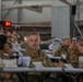 A military briefing or conference is taking place in what appears to be a large industrial or hangar-style facility. Multiple military personnel in camouflage uniforms, along with some civilians in business attire, are seated at long tables with documents, microphones, and beverages. The central figure, a senior military officer wearing glasses, appears to be speaking or engaged in discussion.