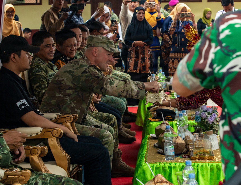 A military ceremony or cultural event appears to be taking place, likely in Indonesia, where personnel in various military uniforms are seated as honored guests. A US soldier in MultiCam/OCP uniform is receiving something from a person in traditional ceremonial attire. The event is being photographed by numerous attendees in the background, suggesting a formal bilateral or multinational military engagement.