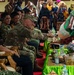 A military ceremony or cultural event appears to be taking place, likely in Indonesia, where personnel in various military uniforms are seated as honored guests. A US soldier in MultiCam/OCP uniform is receiving something from a person in traditional ceremonial attire. The event is being photographed by numerous attendees in the background, suggesting a formal bilateral or multinational military engagement.