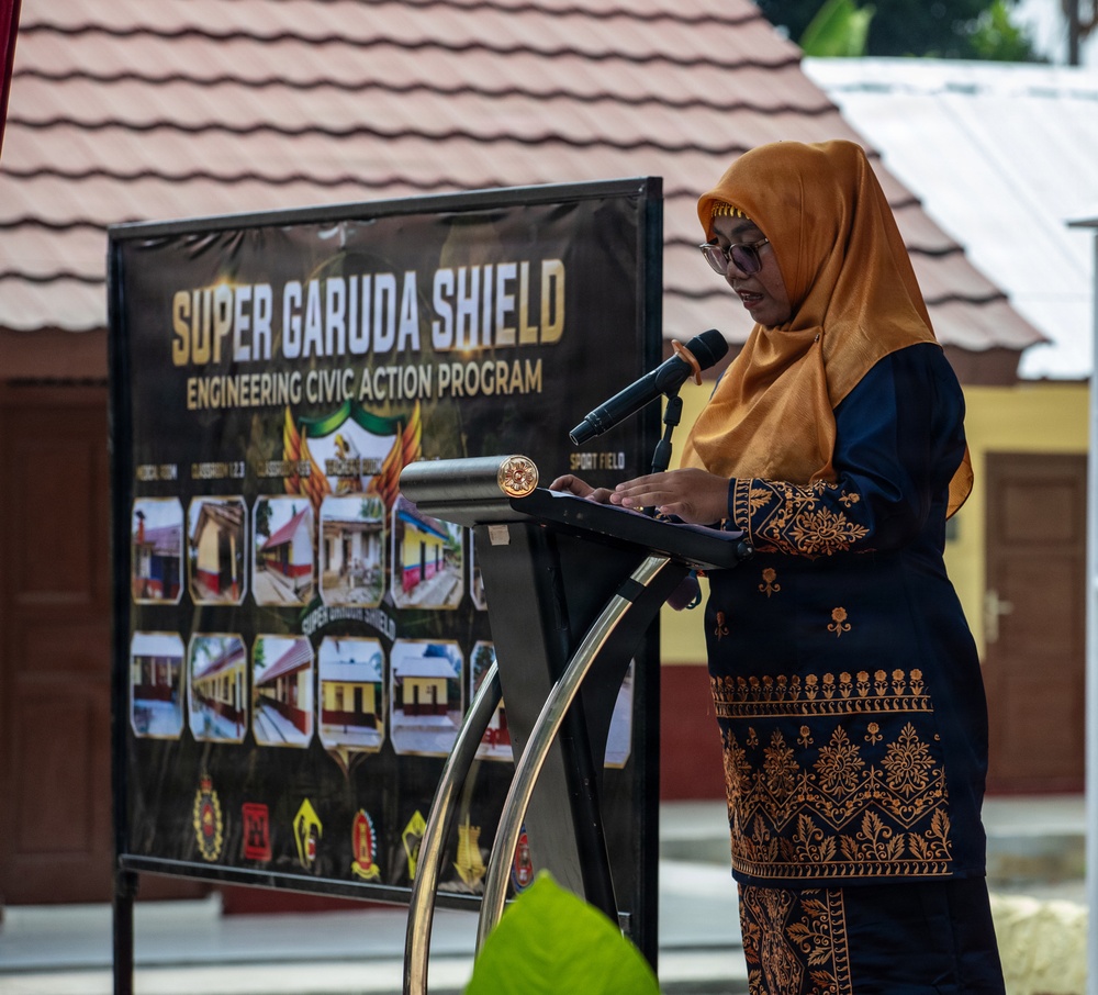 A woman wearing a golden hijab and dark blue batik attire is speaking at a podium with a microphone during an official event. Behind her is a banner reading 'Super Garuda Shield - Engineering Civic Action Program' featuring photographs of various constructed facilities including classrooms and sports fields. The event appears to be a military-civic cooperation program ceremony, likely held in Indonesia.