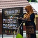 A woman wearing a golden hijab and dark blue batik attire is speaking at a podium with a microphone during an official event. Behind her is a banner reading 'Super Garuda Shield - Engineering Civic Action Program' featuring photographs of various constructed facilities including classrooms and sports fields. The event appears to be a military-civic cooperation program ceremony, likely held in Indonesia.