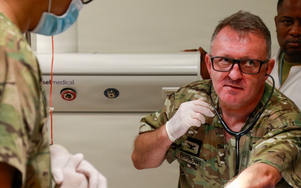 Military medical personnel are treating a patient in what appears to be a field or forward medical facility. A central figure wearing camouflage uniform with a name tape reading 'BURROWS' is using a stethoscope while wearing gloves, with at least two other individuals assisting. The scene depicts an active medical intervention with IV lines and medical equipment visible.