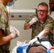 Military medical personnel are treating a patient in what appears to be a field or forward medical facility. A central figure wearing camouflage uniform with a name tape reading 'BURROWS' is using a stethoscope while wearing gloves, with at least two other individuals assisting. The scene depicts an active medical intervention with IV lines and medical equipment visible.