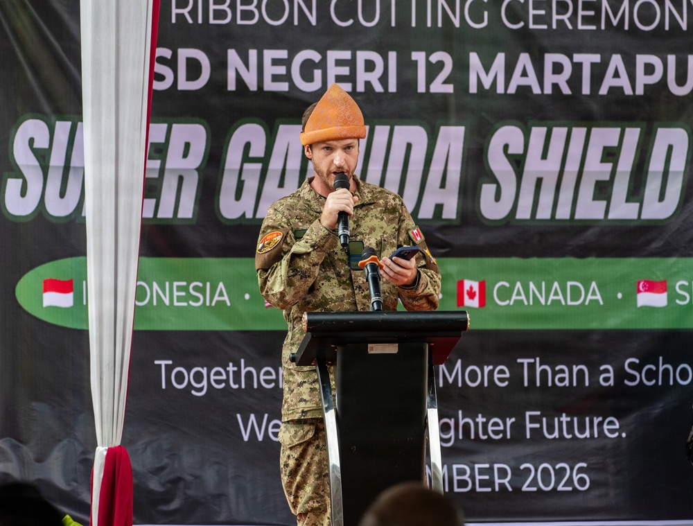 A military personnel member in camouflage uniform is speaking at a podium during a ribbon cutting ceremony for SD Negeri 12 Martapu, associated with the 'Super Garuda Shield' exercise. The speaker is wearing an orange traditional Indonesian hat (blangkon or similar) and holding a microphone and mobile phone. The banner behind indicates participation from Indonesia, Canada, and Singapore, with a date referencing 2026.