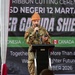 A military personnel member in camouflage uniform is speaking at a podium during a ribbon cutting ceremony for SD Negeri 12 Martapu, associated with the 'Super Garuda Shield' exercise. The speaker is wearing an orange traditional Indonesian hat (blangkon or similar) and holding a microphone and mobile phone. The banner behind indicates participation from Indonesia, Canada, and Singapore, with a date referencing 2026.