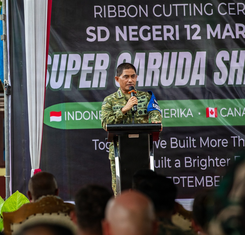 A military officer in camouflage uniform is speaking at a podium during a ribbon cutting ceremony for SD Negeri 12, associated with the 'Super Garuda Shield' exercise. The banner behind him references Indonesia, Amerika (America), and Canada as participating nations. The event appears to be a joint military humanitarian/community project involving the construction or renovation of a school.