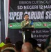 A military officer in camouflage uniform is speaking at a podium during a ribbon cutting ceremony for SD Negeri 12, associated with the 'Super Garuda Shield' exercise. The banner behind him references Indonesia, Amerika (America), and Canada as participating nations. The event appears to be a joint military humanitarian/community project involving the construction or renovation of a school.
