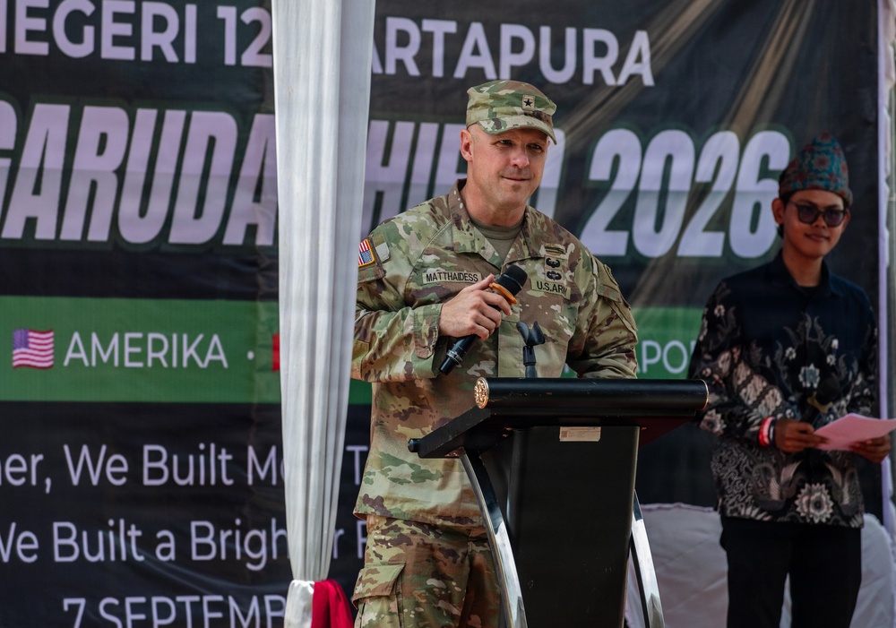 A U.S. Army officer with the name tag 'MATTHAIDESS' and a single star insignia (Brigadier General rank) is speaking at a podium during the 'Garuda Shield 2026' military exercise event. The event appears to be held in Indonesia, indicated by the banner referencing 'Amerika' and the Indonesian text. A civilian in traditional Indonesian attire stands to the right holding a red folder.
