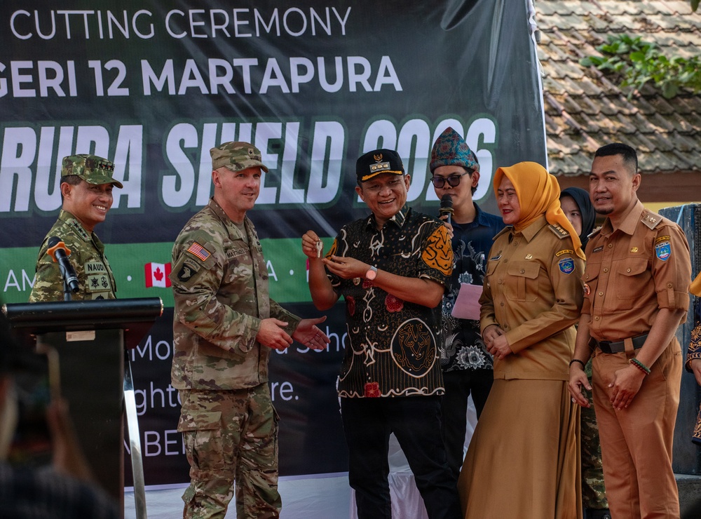 A ribbon-cutting ceremony is taking place at SMP Negeri 12 Martapura as part of the 'Garuda Shield 2023' military exercise. A U.S. Army soldier wearing a 101st Airborne Division patch is participating alongside Indonesian civilian officials and military personnel. The event appears to be a community engagement or civic action project involving U.S. and Indonesian cooperation.
