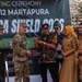 A ribbon-cutting ceremony is taking place at SMP Negeri 12 Martapura as part of the 'Garuda Shield 2023' military exercise. A U.S. Army soldier wearing a 101st Airborne Division patch is participating alongside Indonesian civilian officials and military personnel. The event appears to be a community engagement or civic action project involving U.S. and Indonesian cooperation.