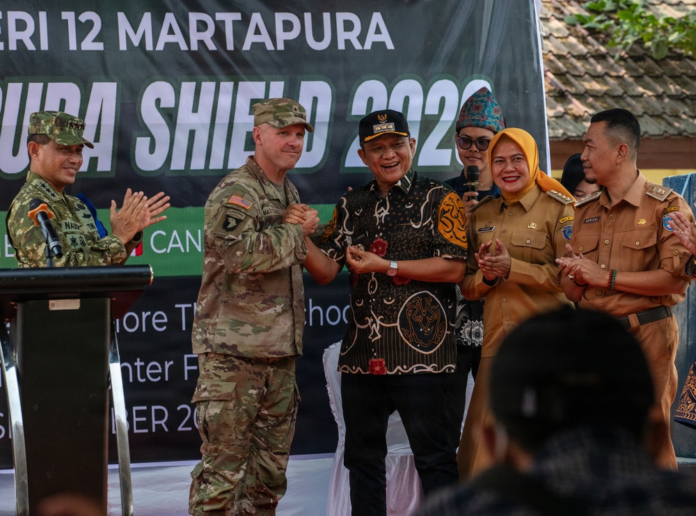 A ceremonial event at SMP Negeri 12 Martapura related to 'Garuda Shield 2023', a joint military exercise. A U.S. Army soldier wearing a 101st Airborne Division patch is shaking hands with a local official in traditional Indonesian attire. The gathering includes Indonesian military, civilian officials, and local dignitaries in a celebratory atmosphere.