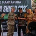 A ceremonial event at SMP Negeri 12 Martapura related to 'Garuda Shield 2023', a joint military exercise. A U.S. Army soldier wearing a 101st Airborne Division patch is shaking hands with a local official in traditional Indonesian attire. The gathering includes Indonesian military, civilian officials, and local dignitaries in a celebratory atmosphere.