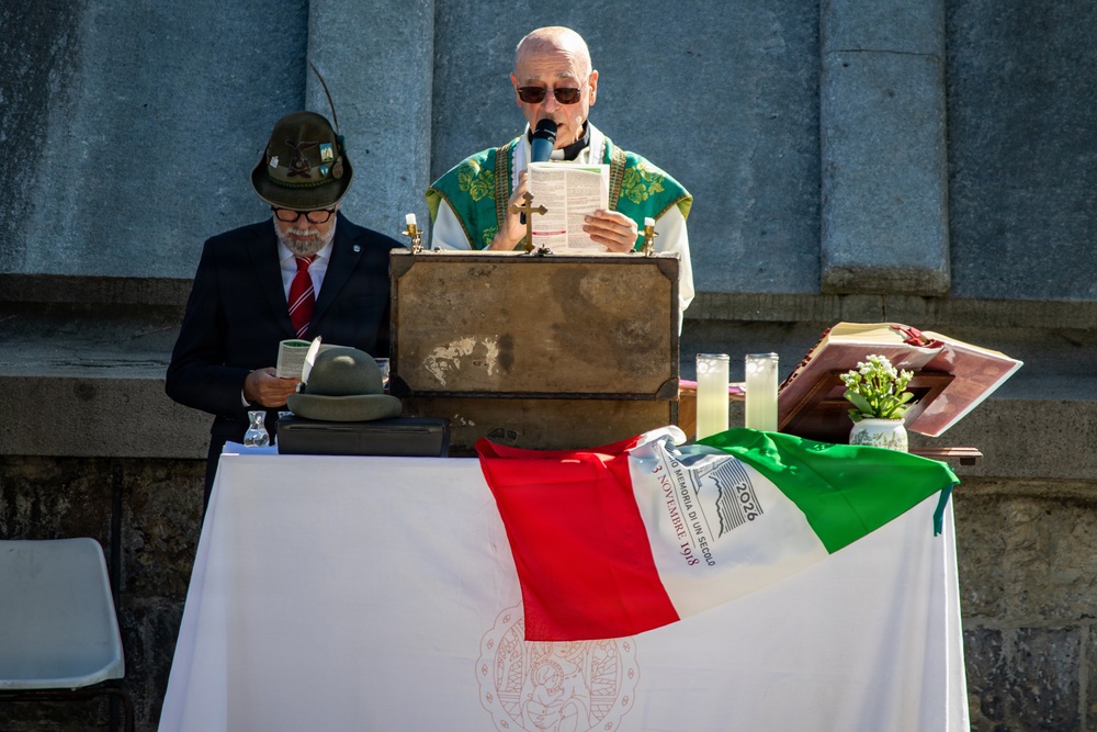 An outdoor Catholic religious ceremony is taking place, likely a commemorative Mass. A priest in green liturgical vestments reads from a document at a podium, while a man in a suit wearing a traditional Alpine hat (cappello alpino) stands nearby. An Italian flag and what appears to be an Alpini (Italian mountain troops) commemorative banner are draped over the altar table.