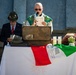 An outdoor Catholic religious ceremony is taking place, likely a commemorative Mass. A priest in green liturgical vestments reads from a document at a podium, while a man in a suit wearing a traditional Alpine hat (cappello alpino) stands nearby. An Italian flag and what appears to be an Alpini (Italian mountain troops) commemorative banner are draped over the altar table.