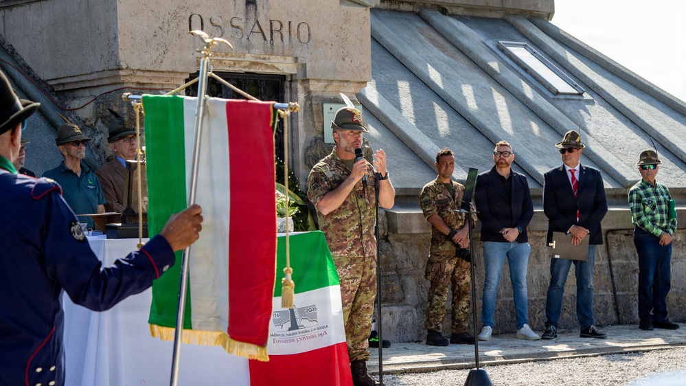 A formal military and civic ceremony is taking place in front of a stone building marked 'OSSARIO' (ossuary/charnel house), likely a war memorial in Italy. A soldier in camouflage uniform is speaking at a microphone while an Italian flag is prominently displayed in the foreground. Several attendees in military uniforms, civilian clothes, and traditional Alpine hats are present.