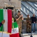 A formal military and civic ceremony is taking place in front of a stone building marked 'OSSARIO' (ossuary/charnel house), likely a war memorial in Italy. A soldier in camouflage uniform is speaking at a microphone while an Italian flag is prominently displayed in the foreground. Several attendees in military uniforms, civilian clothes, and traditional Alpine hats are present.