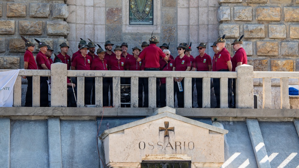 A group of approximately 20 individuals dressed in matching red polo shirts and traditional Alpine-style hats with feathers stand on a stone balcony above an ossuary (bone repository) entrance marked 'OSSARIO' with a cross. They appear to be a choir or musical ensemble, likely performing or preparing to perform at a memorial or commemorative site. The setting suggests a formal ceremony at what appears to be a WWI or WWII Italian military memorial.