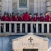A group of approximately 20 individuals dressed in matching red polo shirts and traditional Alpine-style hats with feathers stand on a stone balcony above an ossuary (bone repository) entrance marked 'OSSARIO' with a cross. They appear to be a choir or musical ensemble, likely performing or preparing to perform at a memorial or commemorative site. The setting suggests a formal ceremony at what appears to be a WWI or WWII Italian military memorial.