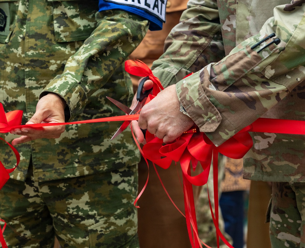 Military personnel in different camouflage uniforms are participating in a ribbon-cutting ceremony. One person holds a red ribbon taut while another cuts it with large scissors, with decorative red bows visible. The image captures a formal ceremonial event, likely marking an opening or inauguration of a military facility or program.