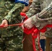 Military personnel in different camouflage uniforms are participating in a ribbon-cutting ceremony. One person holds a red ribbon taut while another cuts it with large scissors, with decorative red bows visible. The image captures a formal ceremonial event, likely marking an opening or inauguration of a military facility or program.