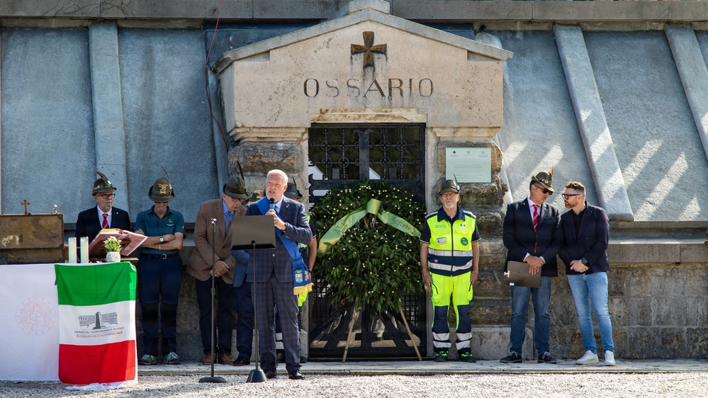 A formal commemorative ceremony is taking place in front of an 'Ossario' (ossuary/charnel house), a structure used to house the remains of the deceased. A speaker addresses attendees at a microphone podium, flanked by officials and participants, with a large floral wreath placed at the entrance. An Italian flag is visible on a table to the left, suggesting this is an official Italian memorial event.