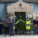 A formal commemorative ceremony is taking place in front of an 'Ossario' (ossuary/charnel house), a structure used to house the remains of the deceased. A speaker addresses attendees at a microphone podium, flanked by officials and participants, with a large floral wreath placed at the entrance. An Italian flag is visible on a table to the left, suggesting this is an official Italian memorial event.