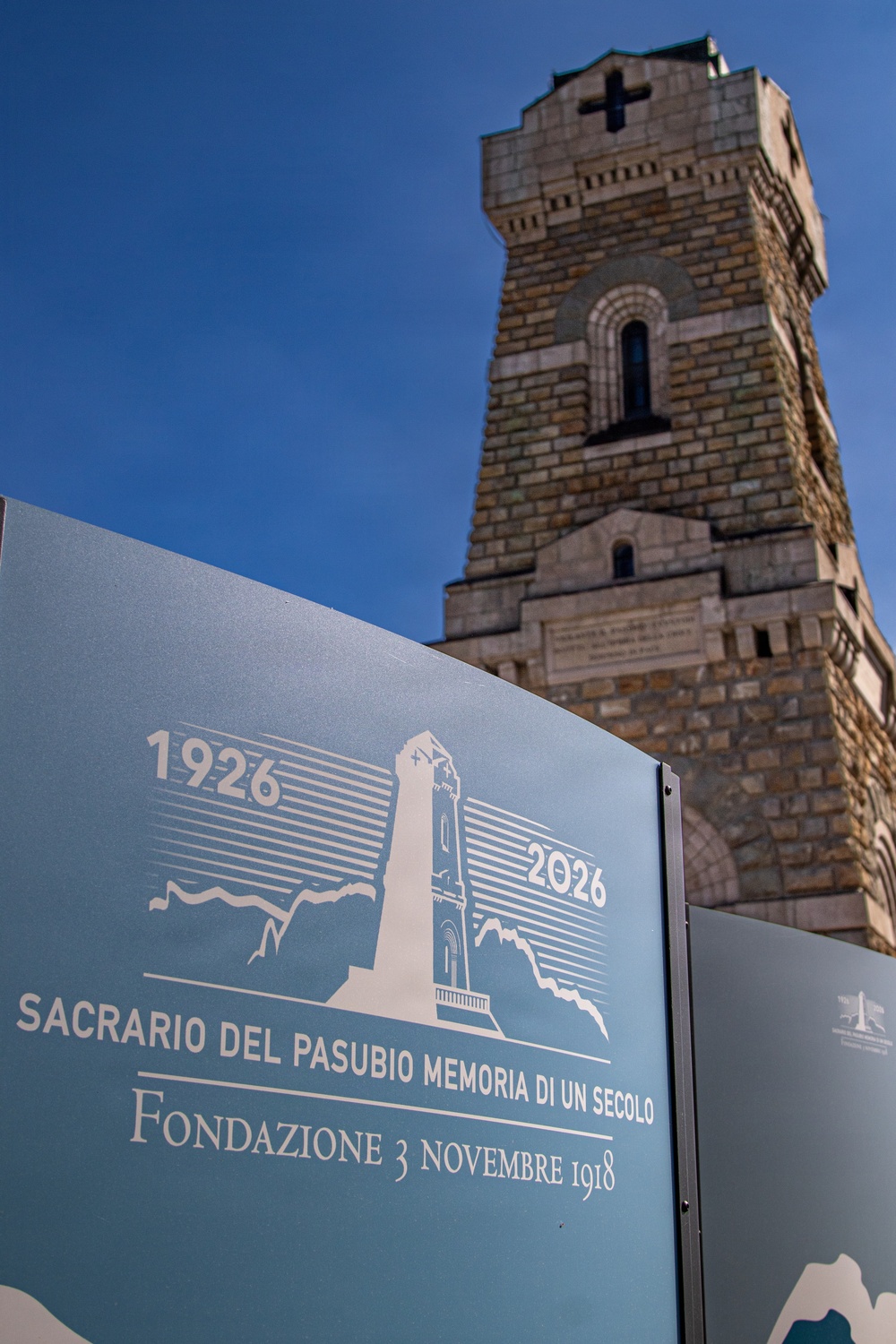 The image shows the Sacrario del Pasubio, a World War I military ossuary and memorial located on Mount Pasubio in northern Italy. In the foreground is a commemorative sign marking the centennial anniversary (1926–2026) of the monument, with the founding date of 3 November 1918. The stone tower with a cross at its summit rises prominently against a clear blue sky in the background.