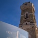 The image shows the Sacrario del Pasubio, a World War I military ossuary and memorial located on Mount Pasubio in northern Italy. In the foreground is a commemorative sign marking the centennial anniversary (1926–2026) of the monument, with the founding date of 3 November 1918. The stone tower with a cross at its summit rises prominently against a clear blue sky in the background.
