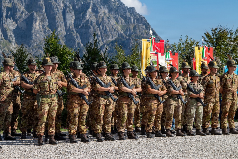 A formation of military personnel in camouflage uniforms and distinctive wide-brimmed alpine hats stand at attention during a formal ceremony. They are armed with assault rifles and positioned in front of colorful regimental or unit flags/banners. The setting appears to be an outdoor parade ground with dramatic rocky mountain peaks visible in the background, consistent with an Italian Alpine troops (Alpini) event.