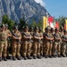 A formation of military personnel in camouflage uniforms and distinctive wide-brimmed alpine hats stand at attention during a formal ceremony. They are armed with assault rifles and positioned in front of colorful regimental or unit flags/banners. The setting appears to be an outdoor parade ground with dramatic rocky mountain peaks visible in the background, consistent with an Italian Alpine troops (Alpini) event.