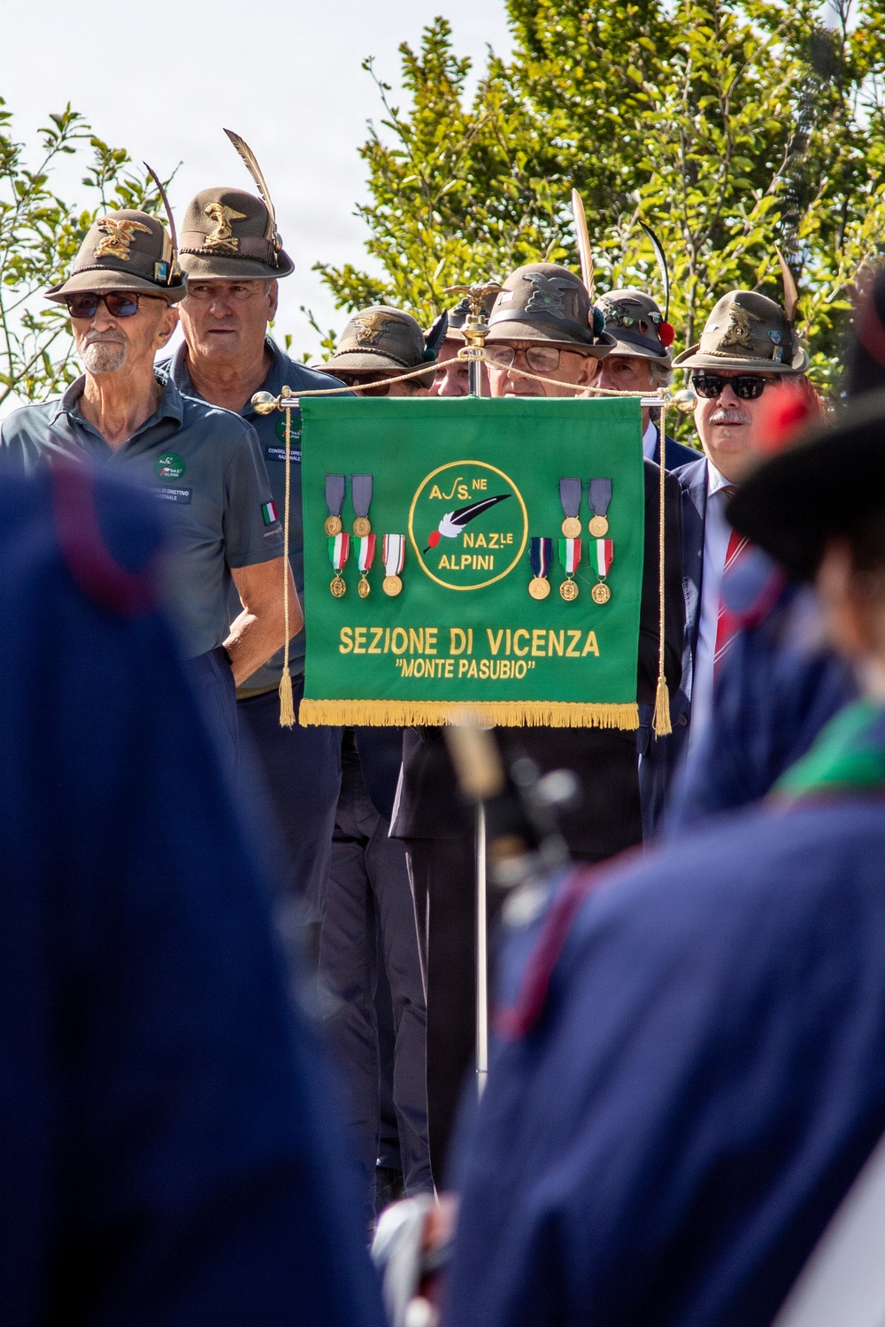 A group of men wearing traditional Alpini hats with feathers are gathered at a formal ceremony. They are displaying a green banner belonging to the 'Associazione Nazionale Alpini - Sezione di Vicenza Monte Pasubio,' decorated with medals and the ANA emblem. The event appears to be an official gathering or parade of the Italian Alpine troops veterans association.