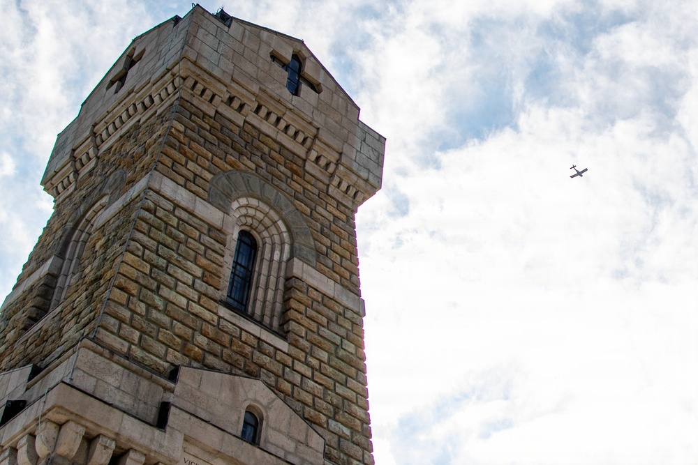 A low-angle upward shot of a large stone tower with Romanesque architectural features, including arched windows and decorative corbeling. The tower is constructed from rough-cut sandstone or limestone blocks. A small aircraft is visible in the partially cloudy sky to the right of the tower.