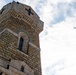 A low-angle upward shot of a large stone tower with Romanesque architectural features, including arched windows and decorative corbeling. The tower is constructed from rough-cut sandstone or limestone blocks. A small aircraft is visible in the partially cloudy sky to the right of the tower.