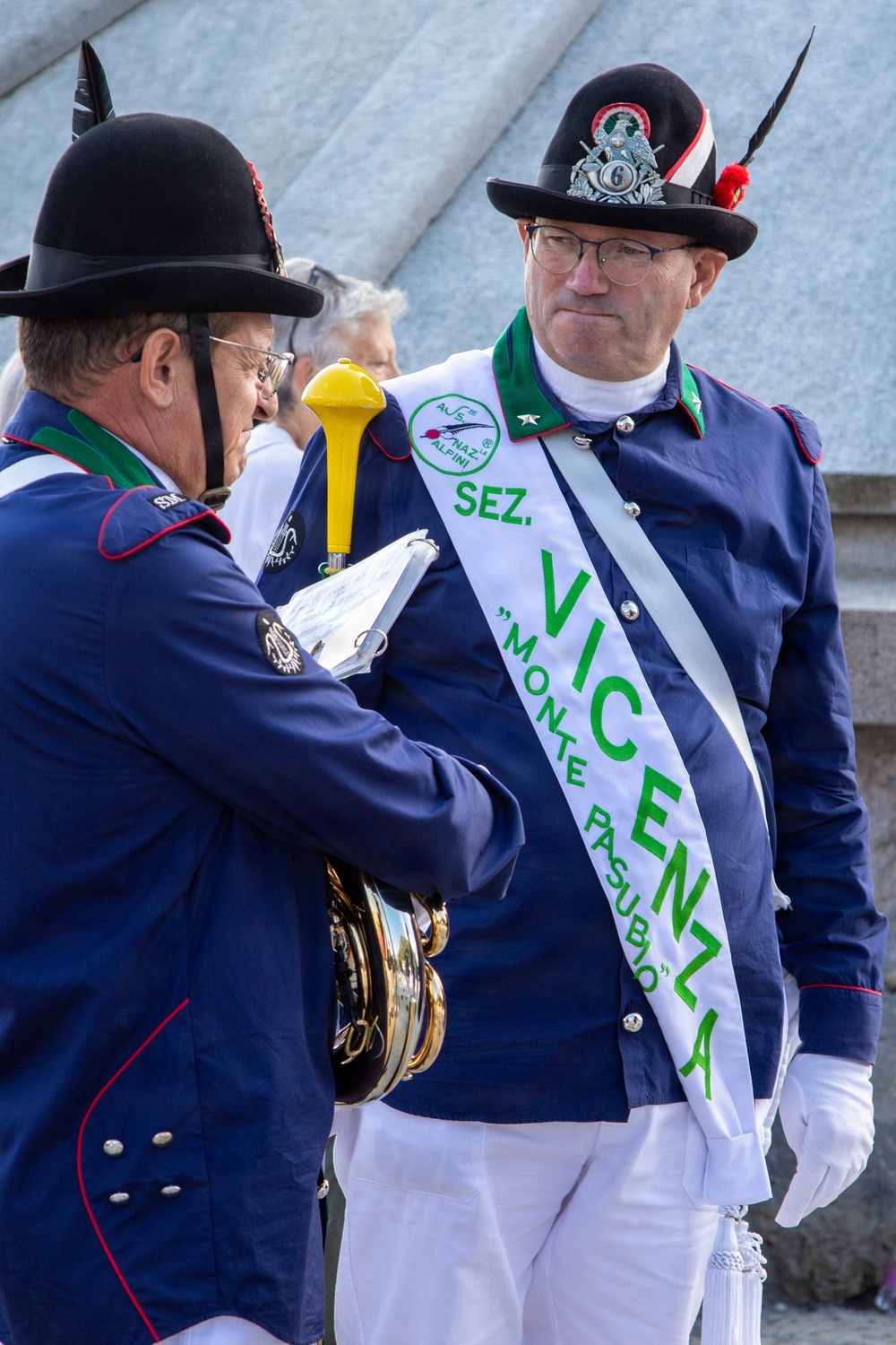 Two men in formal military-style uniforms of the Associazione Nazionale Alpini (Italian Alpine troops veterans association) are engaged in conversation. The man on the right wears a sash identifying him as a representative of the Vicenza section, specifically the 'Monte Pasubio' group. They appear to be at an official Alpini gathering or parade event.