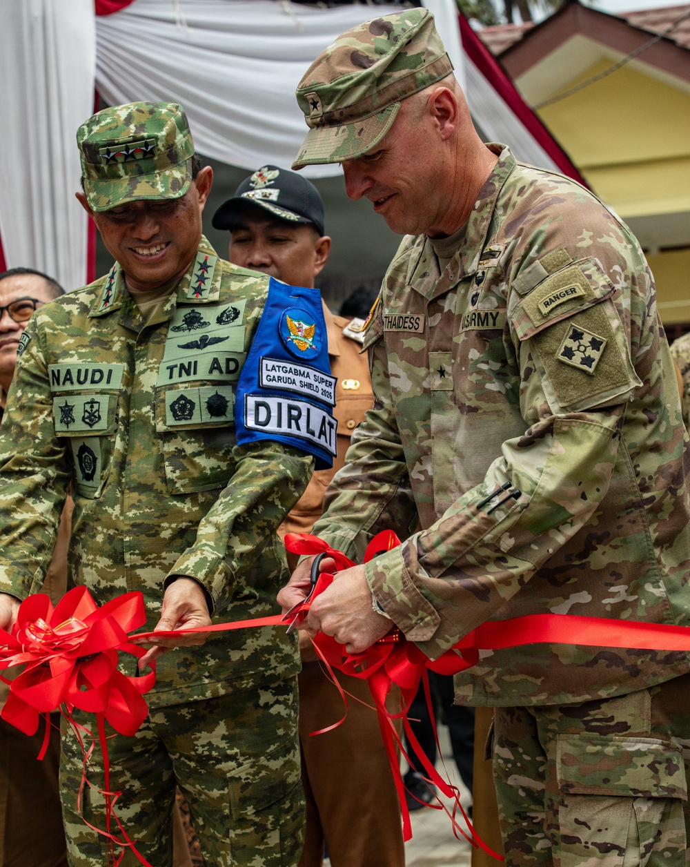 Two military officers, one from the Indonesian Army (TNI AD) and one from the U.S. Army, are jointly cutting a red ribbon at a ceremonial opening event. A third military figure wearing a 'LATGABMA SUPER GARUDA SHIELD 2025 DIRLAT' vest is visible in the background. The event appears to be associated with the Garuda Shield 2025 joint military exercise between Indonesia and the United States.