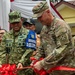 Two military officers, one from the Indonesian Army (TNI AD) and one from the U.S. Army, are jointly cutting a red ribbon at a ceremonial opening event. A third military figure wearing a 'LATGABMA SUPER GARUDA SHIELD 2025 DIRLAT' vest is visible in the background. The event appears to be associated with the Garuda Shield 2025 joint military exercise between Indonesia and the United States.