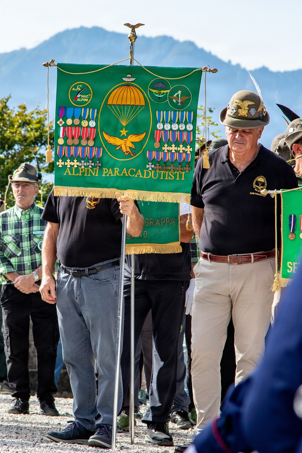 A ceremonial gathering of Italian 'Alpini Paracadutisti' (Alpine Paratroopers) veterans, featuring a prominent green banner adorned with medals, military insignia, a parachute emblem, and an eagle motif. Several men wearing traditional Alpine hats with feathers and black polo shirts bearing unit insignia are present. The setting appears to be an outdoor commemorative event with a mountainous backdrop.