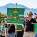 A ceremonial gathering of Italian 'Alpini Paracadutisti' (Alpine Paratroopers) veterans, featuring a prominent green banner adorned with medals, military insignia, a parachute emblem, and an eagle motif. Several men wearing traditional Alpine hats with feathers and black polo shirts bearing unit insignia are present. The setting appears to be an outdoor commemorative event with a mountainous backdrop.
