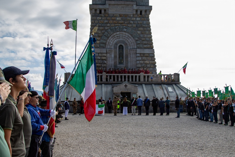 A formal commemorative ceremony is taking place at what appears to be an Italian war memorial ossuary (the word 'OSSARIO' is visible on the structure). Participants are lined up on both sides holding Italian national flags and green organizational banners, facing a stone monument tower. Officials and dignitaries are gathered at the entrance of the memorial, with a choir or honor group visible on the balcony above.