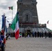 A formal commemorative ceremony is taking place at what appears to be an Italian war memorial ossuary (the word 'OSSARIO' is visible on the structure). Participants are lined up on both sides holding Italian national flags and green organizational banners, facing a stone monument tower. Officials and dignitaries are gathered at the entrance of the memorial, with a choir or honor group visible on the balcony above.