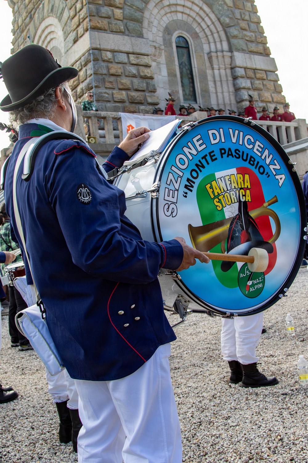 A musician in uniform plays a large bass drum bearing the insignia of the 'Fanfara Storica - Sezione di Vicenza Monte Pasubio,' associated with the Associazione Nazionale Alpini (Italian Alpine troops veterans). The scene takes place outdoors near a stone monument or chapel, with other uniformed participants visible in the background. This appears to be a formal commemorative or ceremonial event.