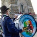 A musician in uniform plays a large bass drum bearing the insignia of the 'Fanfara Storica - Sezione di Vicenza Monte Pasubio,' associated with the Associazione Nazionale Alpini (Italian Alpine troops veterans). The scene takes place outdoors near a stone monument or chapel, with other uniformed participants visible in the background. This appears to be a formal commemorative or ceremonial event.